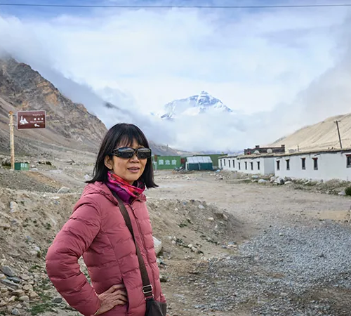 A woman in a pink jacket stands on a gravel path near Rongbuk. Mount Everest appears behind her, a view accessible with an Everest Base Camp Tibet permit.