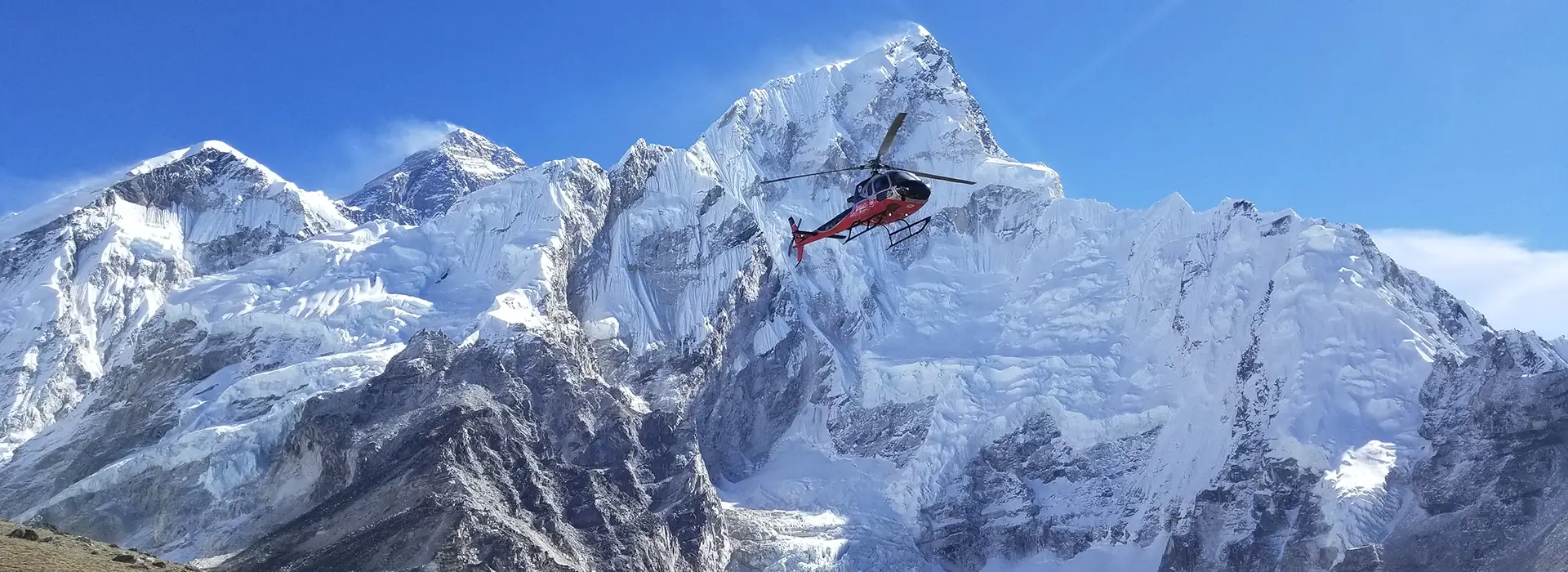 A red and black helicopter flies in a clear blue sky, close to towering, snow-covered Himalayan peaks which are top tourist attractions in Nepal.