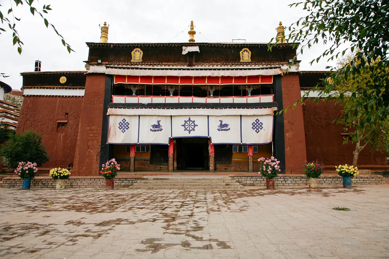 A red stone Tibetan monastery with traditional banners and flower pots in the courtyard. This site is a key cultural stop on a Lhasa to Everest Base Camp Tour.
