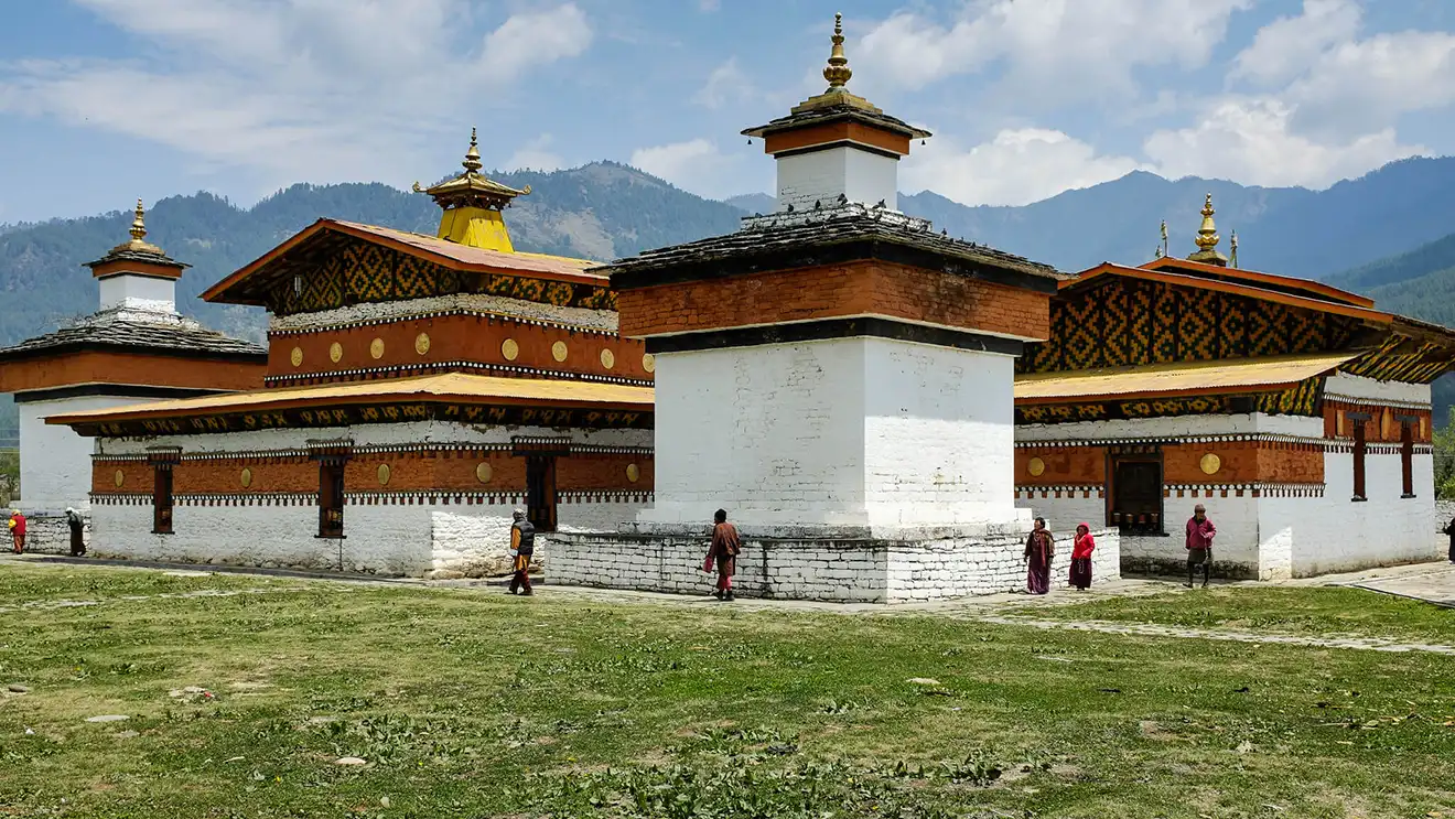 A man wearing a black safety jacket stands next to a parked Royal Enfield adventure motorcycle in front of an ancient Bhutanese temple during a Bhutan Motorcycle Tour.