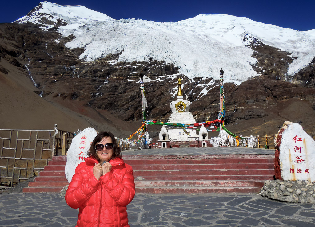Traveler standing in front of Karola Glacier in Tibet, with a white stupa and colorful prayer flags beneath dramatic snow-covered peaks.