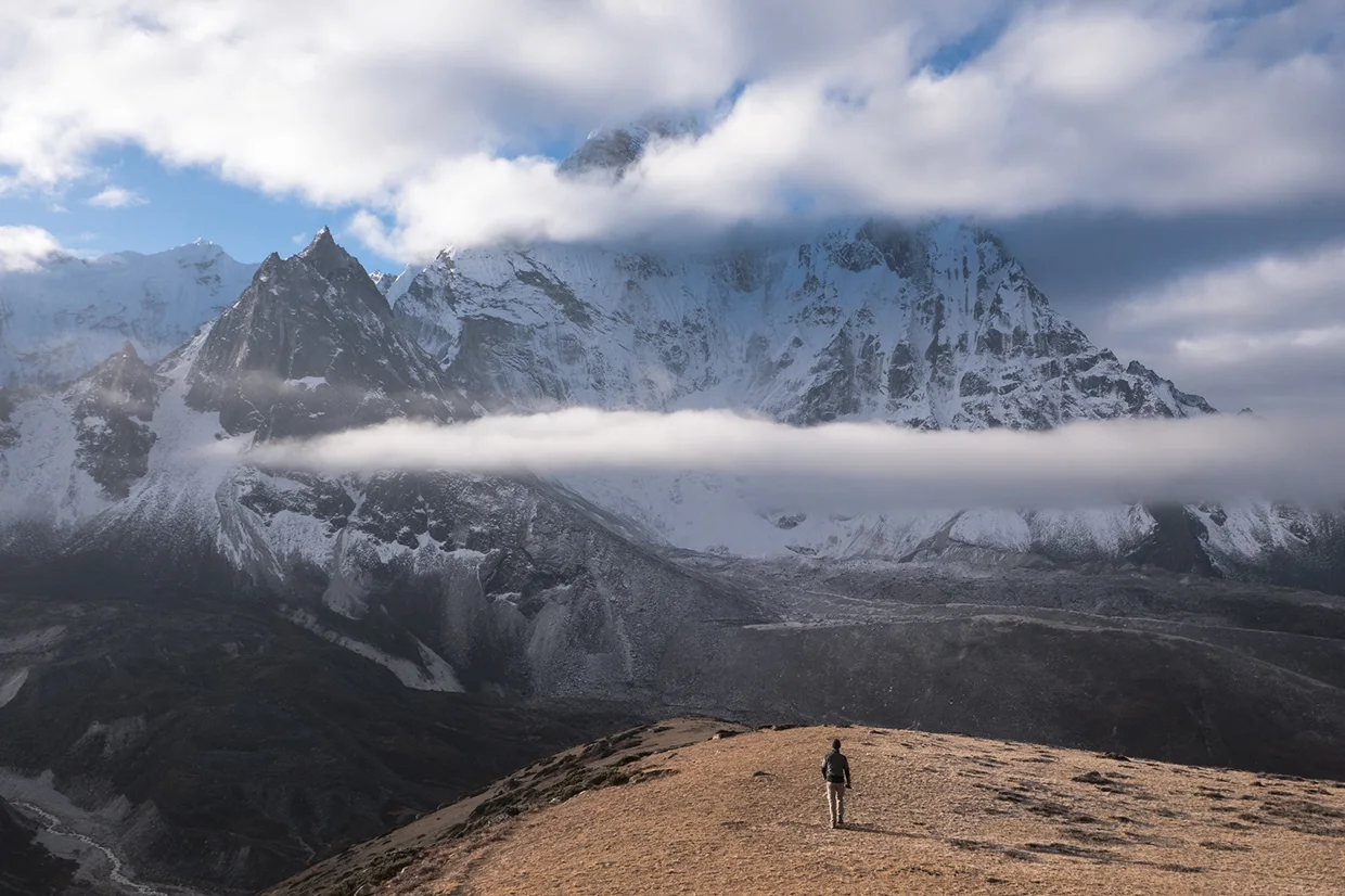 A lone traveler walks along a high mountain ridge toward massive, snow-covered Himalayan peaks partially hidden by clouds during an Everest Three Passes Trek.
