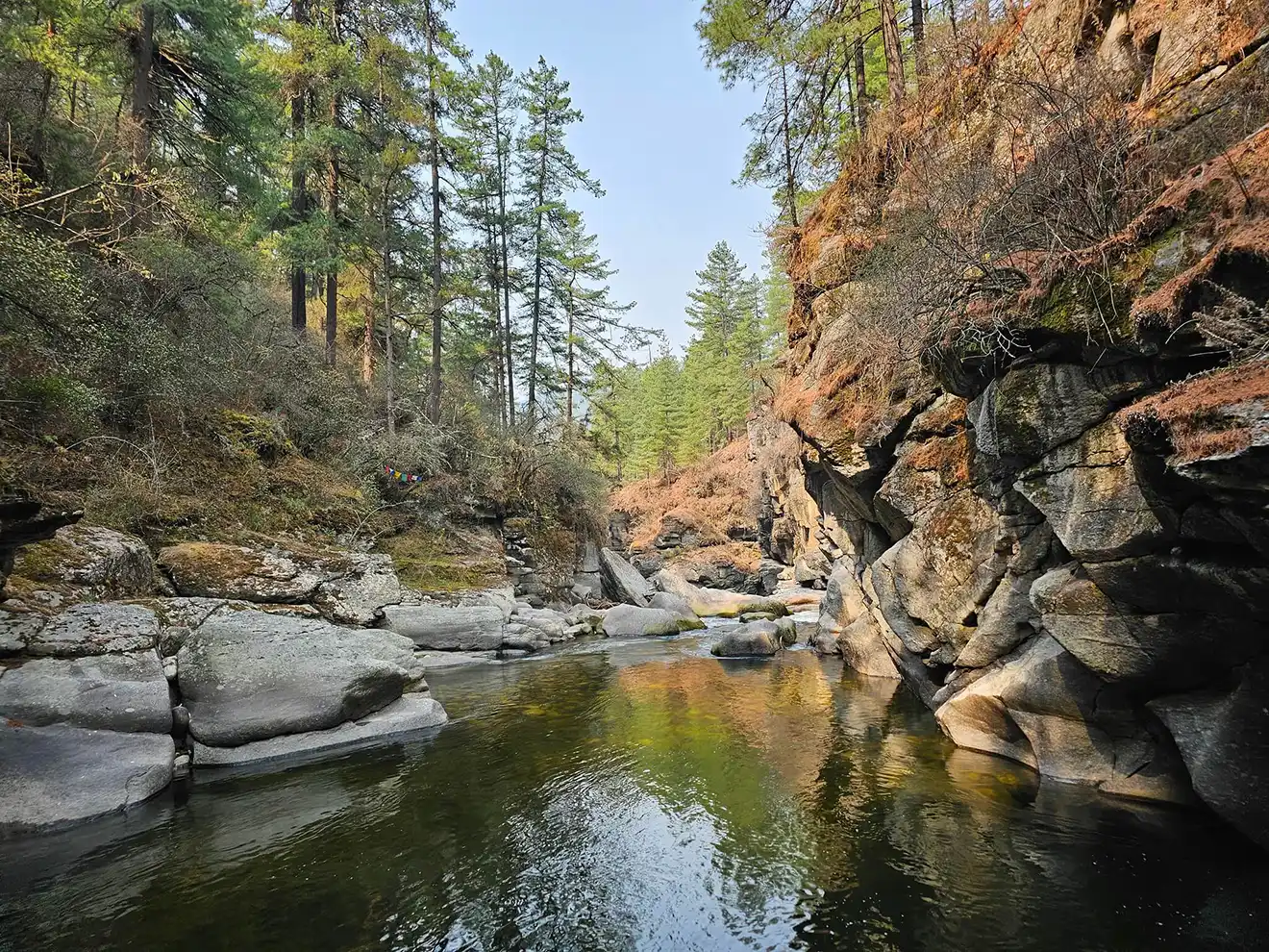 High cliffs and pine trees surround the sacred Mebar Tsho Burning Lake, a significant spiritual site visited on a Bumthang Cultural Trek and Tour.