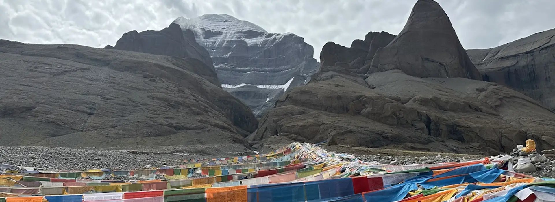 Vibrant Tibetan prayer flags stretch across a rocky valley leading toward the snow-dusted peak of Mount Kailash. Travelers should follow a Mount Kailash packing list for this trek.
