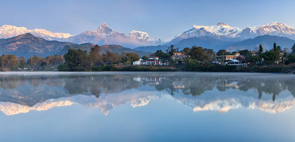 Snow-capped Himalayan peaks rise above Pokhara and reflect clearly in a calm lake, with lakeside houses, trees, and soft morning mist in the foreground.