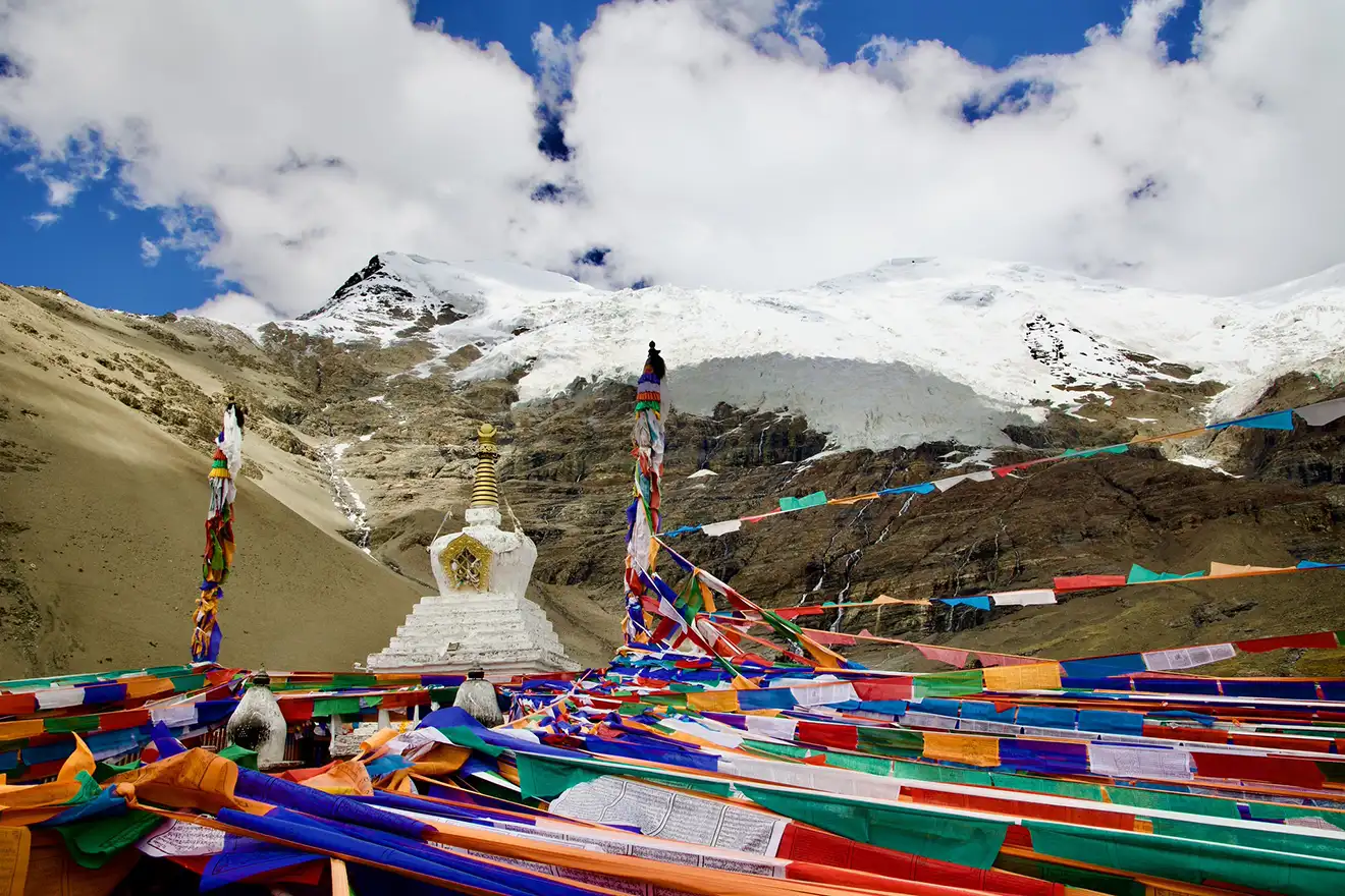 Colorful prayer flags surround a white stone stupa in front of a snow-capped Himalayan peak during a Lhasa to Everest Base Camp Tour.