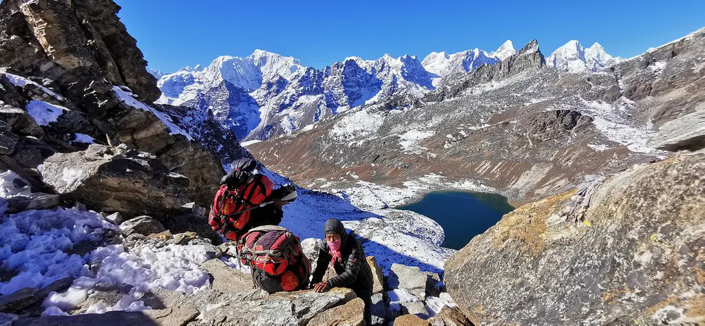 Two local porters carry heavy red gear bags up a rocky, snow-dusted mountain pass overlooking a turquoise alpine lake during an Everest Three Passes Trek.