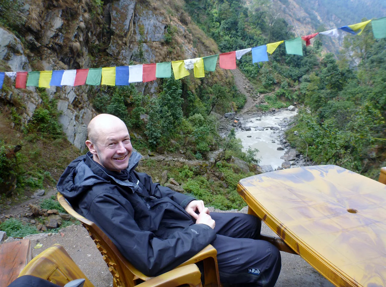 Traveler resting at a riverside stop during the Langtang Trek in Nepal, with prayer flags and a forested mountain valley in the background