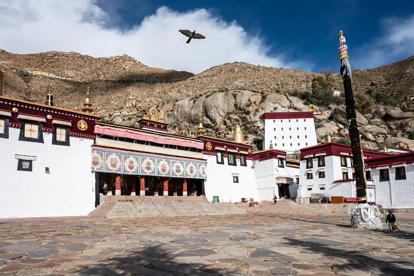 White stone buildings of a Tibetan monastery sit against a rocky mountain backdrop. This peaceful stop serves as a highlight during a Lhasa to Everest Base Camp Tour.