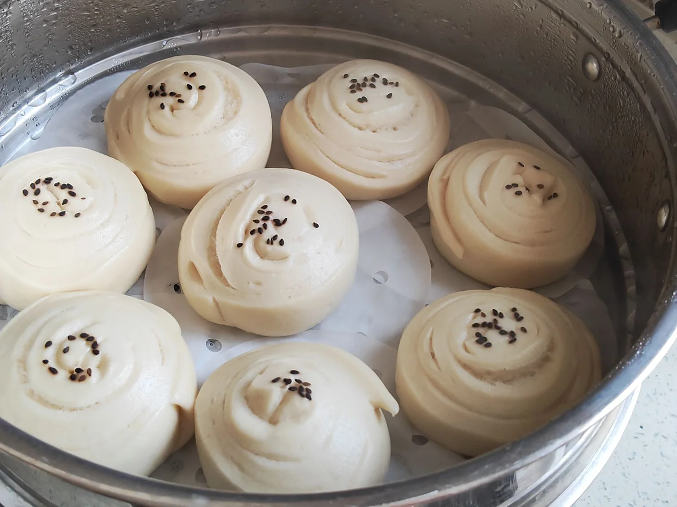 Freshly steamed Tingmo bread rolls with black sesame seeds sit in a metal steamer basket, showcasing a staple side dish in authentic Tibetan food.