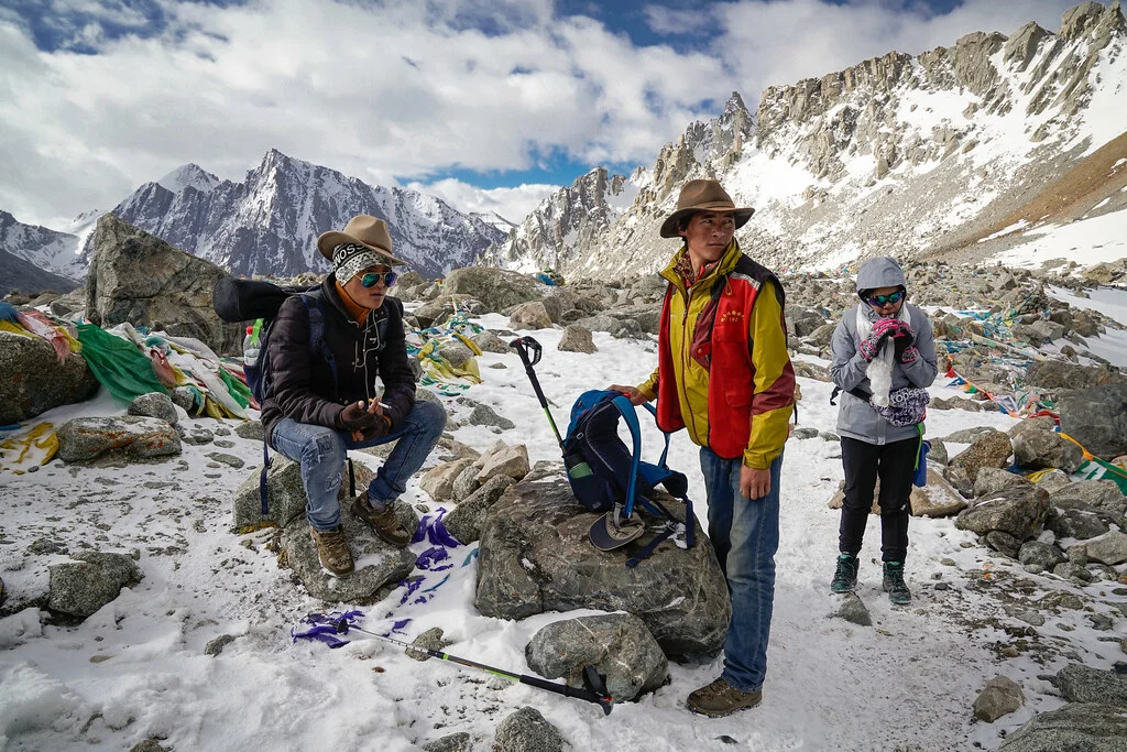 Trekkers resting at Dolma La Pass in Tibet, surrounded by snow-covered mountains, rocky high-altitude terrain, and prayer flags.
