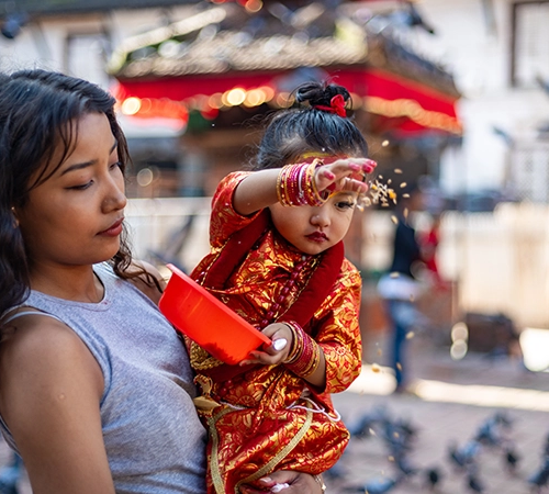A young girl in traditional red Kumari attire throws grain to pigeons in Kathmandu Durbar Square, a cultural highlight for any 7-Day Nepal Itinerary.