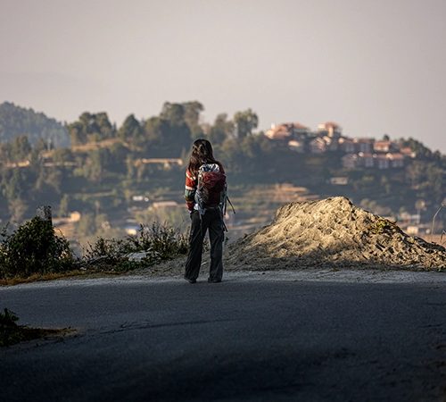 A woman with a backpack stands on a mountain road looking toward a village. This scene captures the exploration of Nepal bucket list destinations.