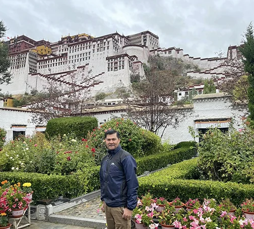 A traveler stands in a lush garden featuring colorful flowers and manicured hedges with the historic Potala Palace rising behind him during a Lhasa and Yamdrok Lake Tour.
