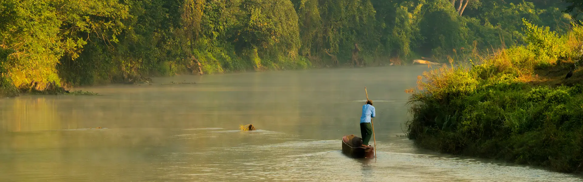 A local man paddles a traditional dugout canoe through the morning mist on the Rapti River, illustrating a serene Chitwan Travel Guide adventure.