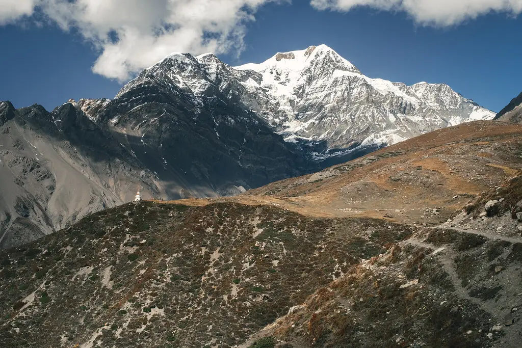 A remote trekking trail winds toward massive snow-capped Himalayan peaks. A hiker can be seen near a small, white chorten along a path that travelers explore during a comprehensive Nepal itinerary for 7 days.