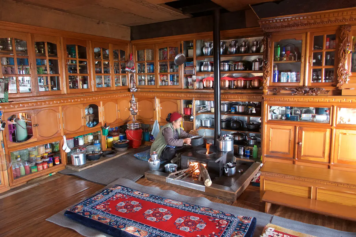 A woman cooks in a traditional Himalayan kitchen with wooden cabinets and a central wood-burning stove, illustrating the cultural items on a Nepal Hiking Packing List.