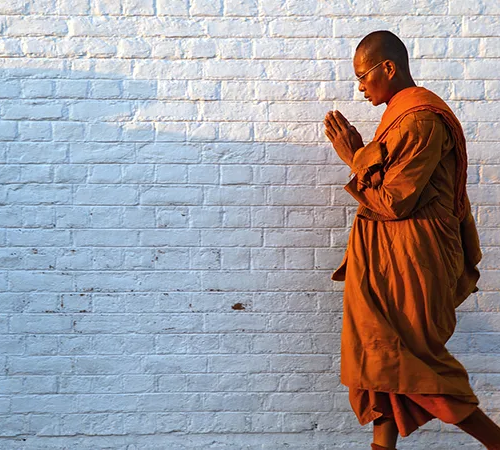 A Theravada Buddhist monk in saffron robes walks with hands clasped in prayer against a white brick wall. This serene moment is a highlight of this Lumbini Travel Guide.