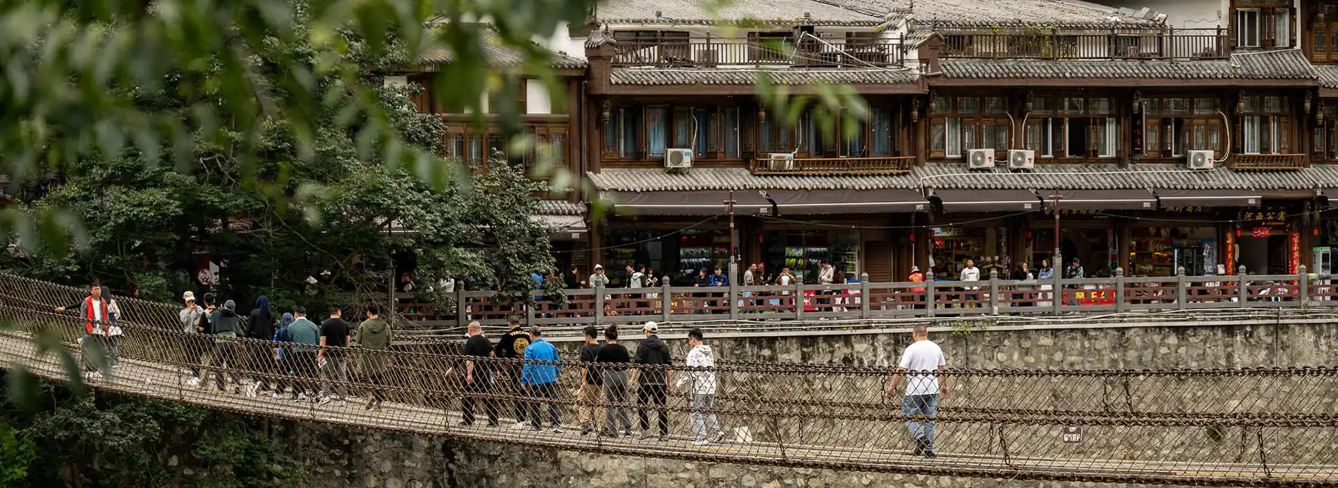 Pedestrians cross a narrow suspension bridge toward a traditional town with wooden buildings. Travelers visiting sensitive border towns must carry an official Tibet Military Permit.