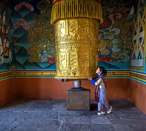 A young Bhutanese girl in traditional dress spins a large golden prayer wheel inside a colorful temple during an authentic Bhutan Cultural Tour Package.