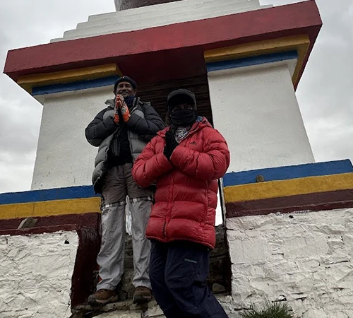 Two travelers in down jackets pose with prayer hands in front of a colorful traditional Tibetan stupa. This scenic stop is part of our Kailash Tour from Lhasa.