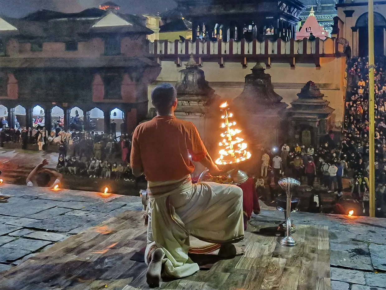 A large crowd gathers on the riverbank at dusk, holding lit butter lamps and watching an evening aarti ceremony, one of the essential spiritual things to do in Kathmandu.