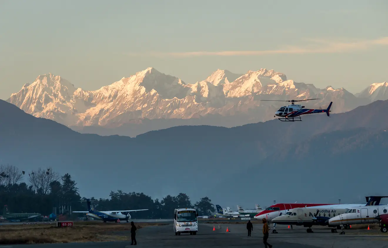 A helicopter takes off from a busy airport tarmac, with several airplanes visible. The majestic snow-capped Himalayan peaks loom in the background under a clear sky, a key view for any 7-Day Nepal Itinerary.