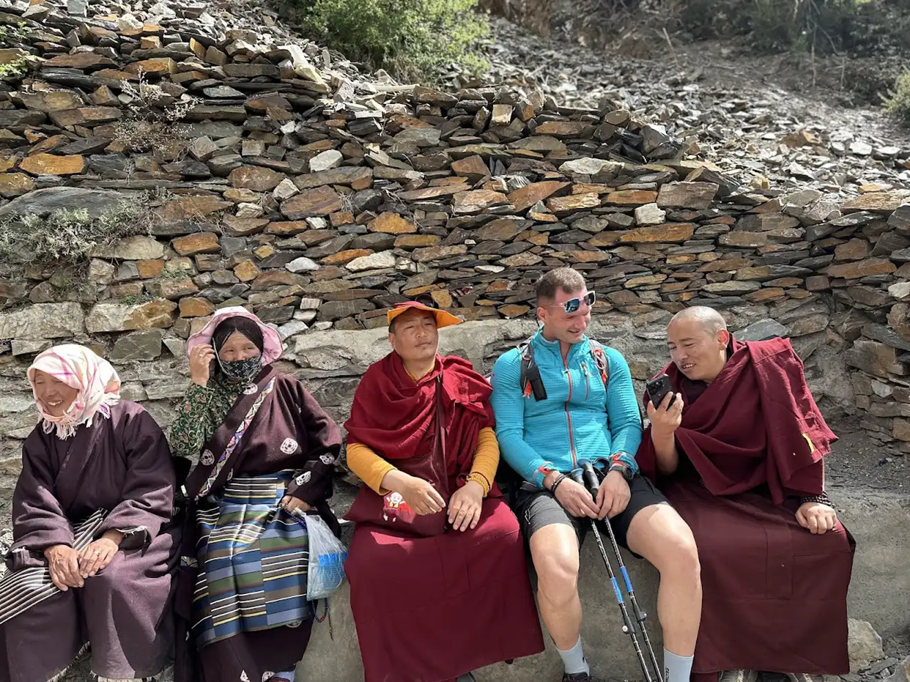 A male hiker sits with Tibetan monks and local women against a stone wall. His technical outdoor gear highlights essential items from a Tibet packing list.