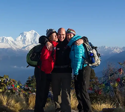 Three happy hikers embrace on a mountain ridge with the Himalayas behind them. Understanding the Nepal Weather by Month helps travelers pick the best dates for clear views.