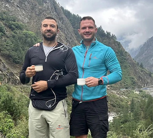 Two male hikers hold tea mugs while standing in a lush Tibetan mountain valley. Specialized travel documents like a Tibet Military Permit are required to visit these restricted zones.