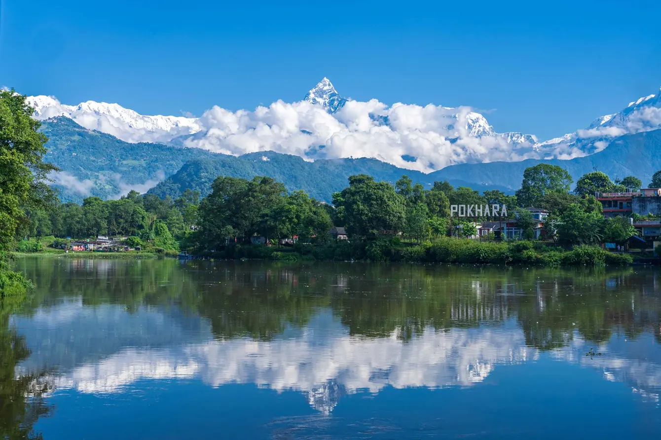 A wide landscape photograph of Phewa Lake in Nepal, showing lush green hills, snow-capped mountains, and a clear blue sky, one of the best places to visit in Pokhara.