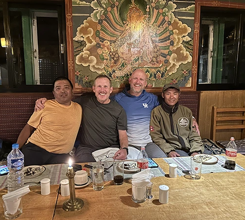 Four men sit together at a wooden table inside a cozy mountain lodge. Traditional Buddhist art appears on the wall behind this top tourist attraction in Nepal.