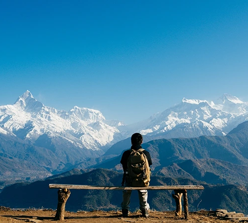 A high-resolution landscape photo from behind a hiker sitting on a rustic bench looking towards the snow-capped Machhapuchhre and Annapurna mountains, illustrating the perfect Pokhara Travel Guide experience.