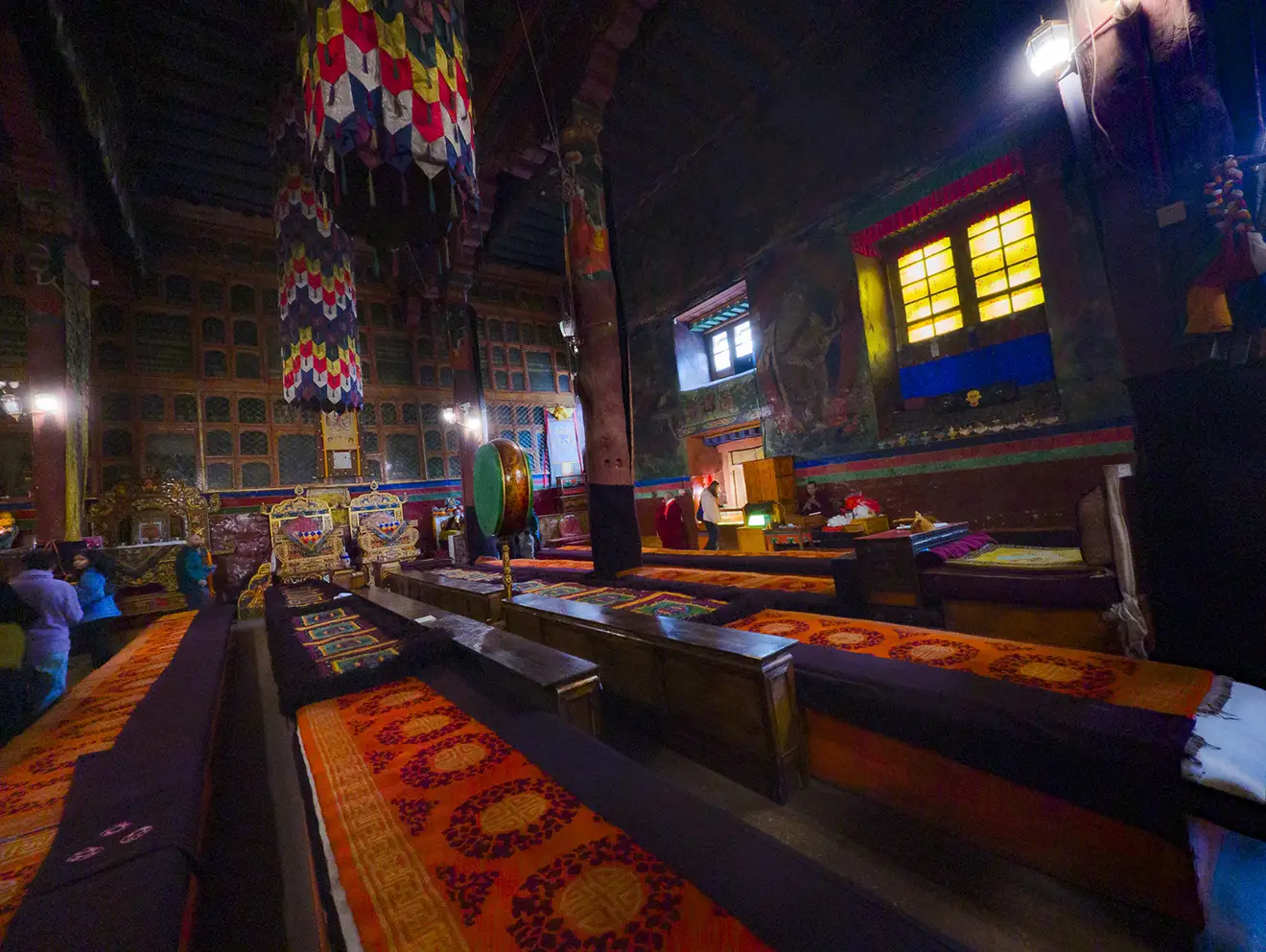 The dimly lit, richly decorated interior of a Tibetan monastery assembly hall, featuring long low benches, hanging textiles, and ancient wall murals, a highlight of a Lhasa to Everest Base Camp and Namtso Lake Tour.