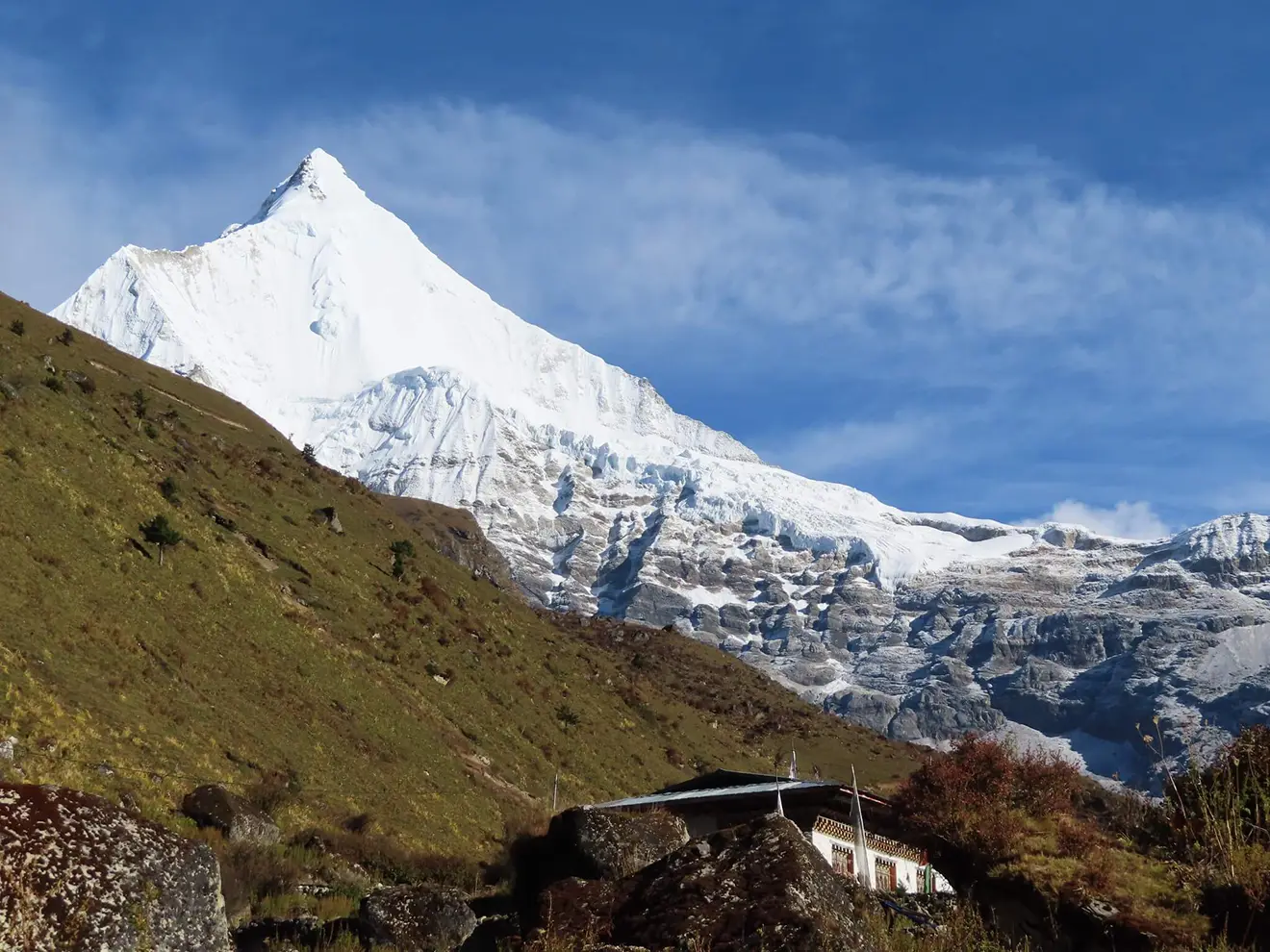 A sharp, snow-covered mountain peak towers over a grassy hillside and a traditional Bhutanese building, showcasing the high-altitude scenery of the Druk Path Trek.