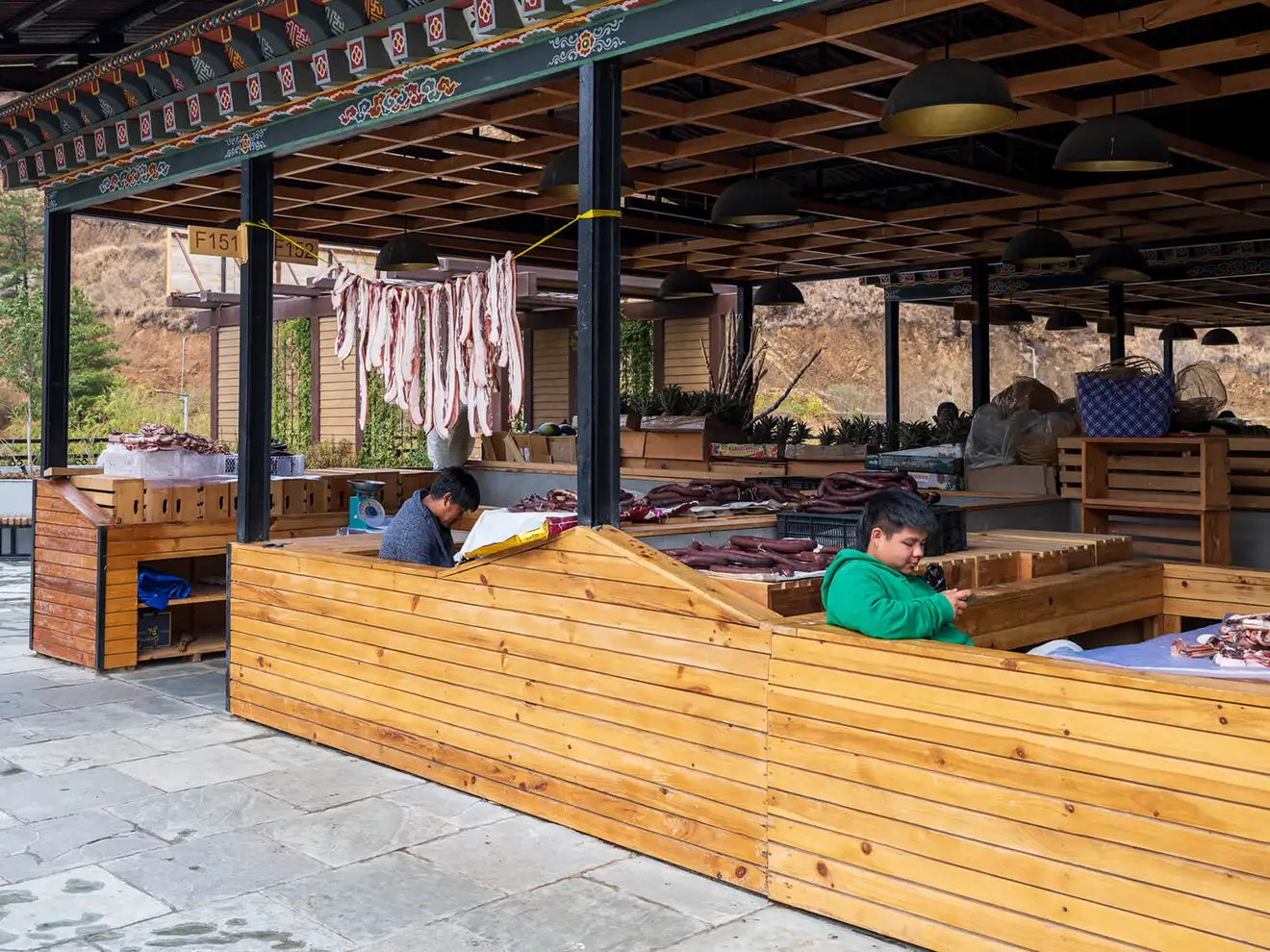 Vendors sit at a local market stall featuring hanging dried meats and fresh produce, a cultural stop during a professional Bhutan Botanical Tour.