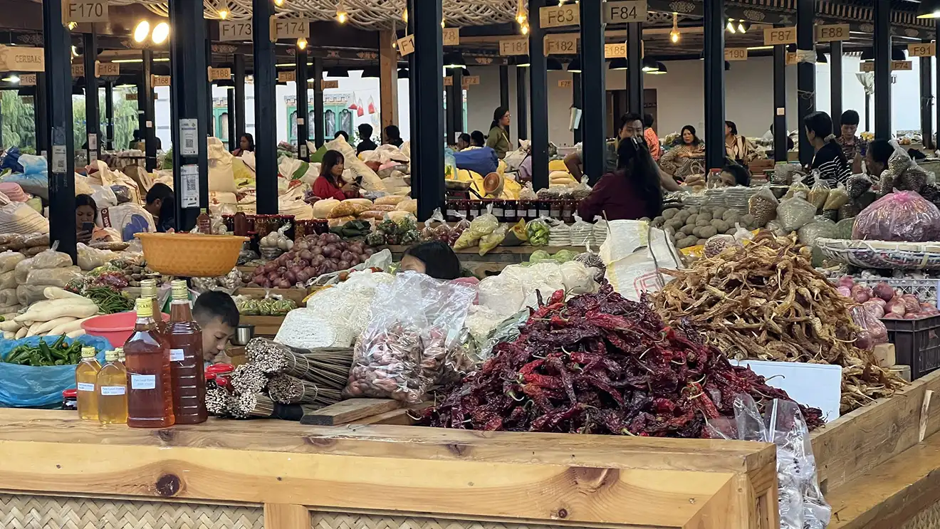 A vibrant, bustling indoor vegetable market in Paro. A central pile of dried red chilies, various bottles of local honey, and produce displays fill the wooden stalls. Local people browse among the vendors, capturing an authentic moment during a Western Bhutan Exploration.