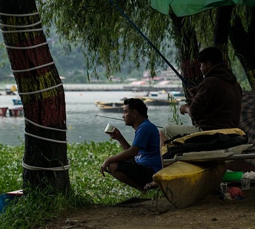 Two local men rest by Phewa Lake, one drinking tea while fishing, showcasing the authentic lifestyle at the best places to visit in Pokhara.