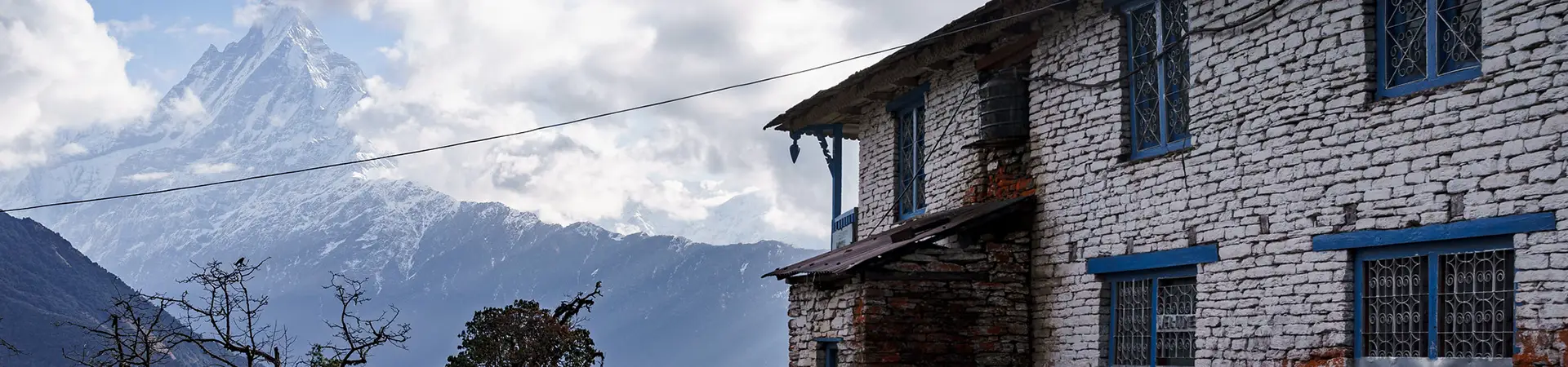 A traditional stone house with blue window frames stands before the towering, snow-covered peak of Machhapuchhre, a view common on a 2-week Nepal itinerary.
