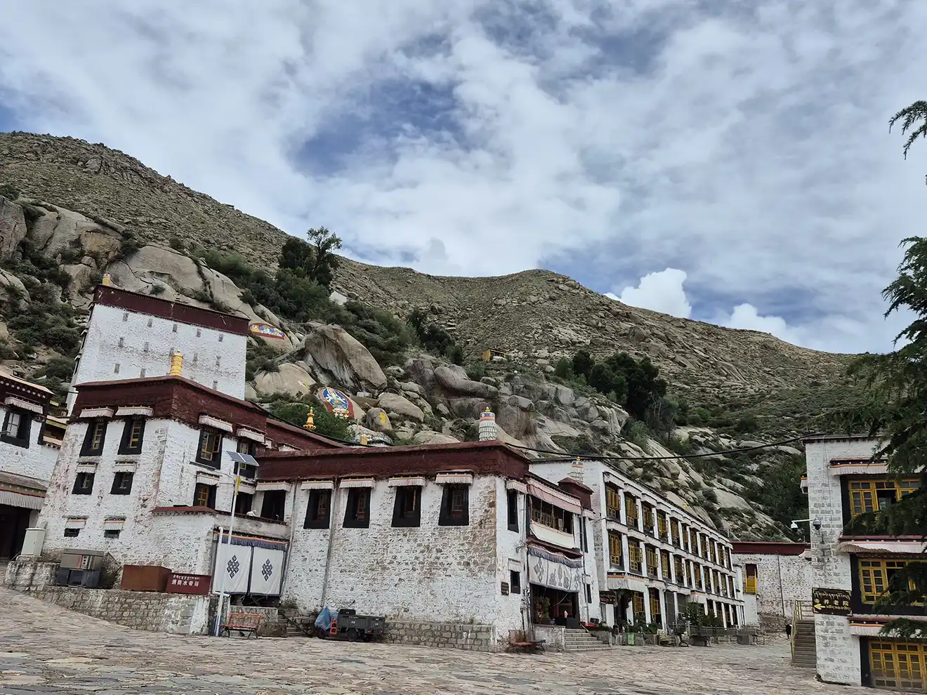 White traditional Tibetan buildings with maroon trim stand against a steep, rocky hill under a cloudy sky, visited on a Lhasa to Kathmandu Overland Tour.