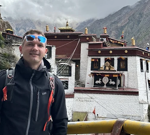 A male trekker in a black jacket and sunglasses stands before a white and red Tibetan monastery. He carries gear from his Mount Kailash packing list.