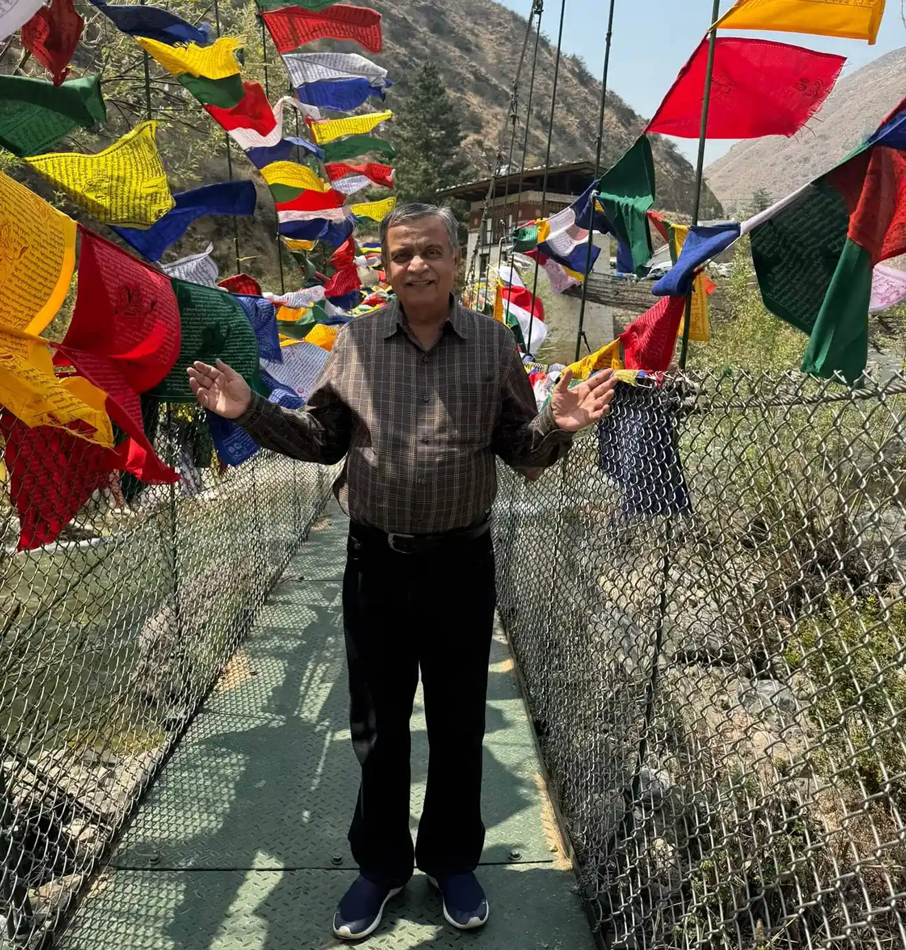 A traveler stands on the historic Tachog Lhakhang iron chain bridge decorated with colorful prayer flags during a guided Bhutan Cultural Tour.