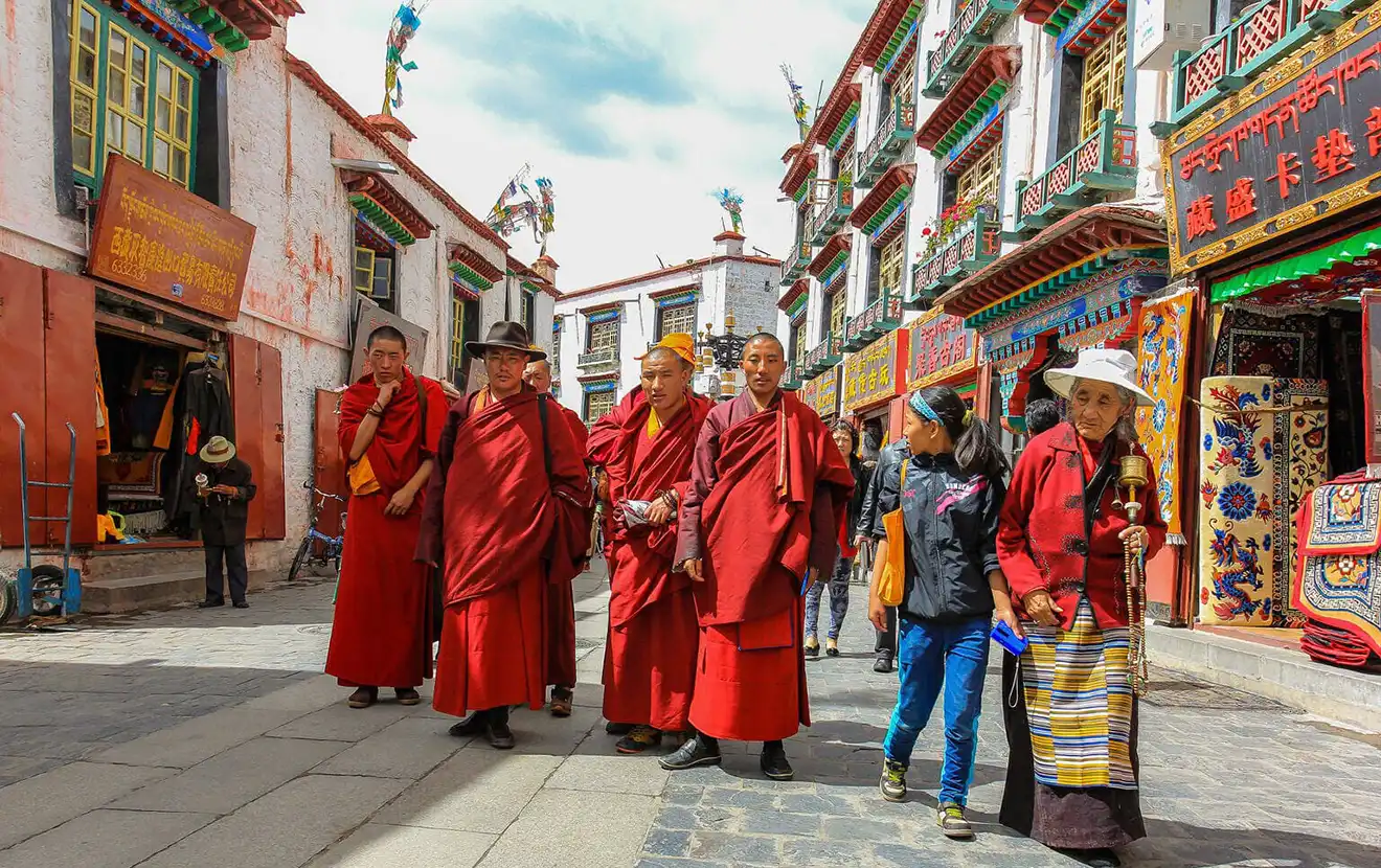 Tibetan monks in red robes walk through a crowded market street lined with shops. Barkhor Street is a prime location for deciding what to buy in Tibet.