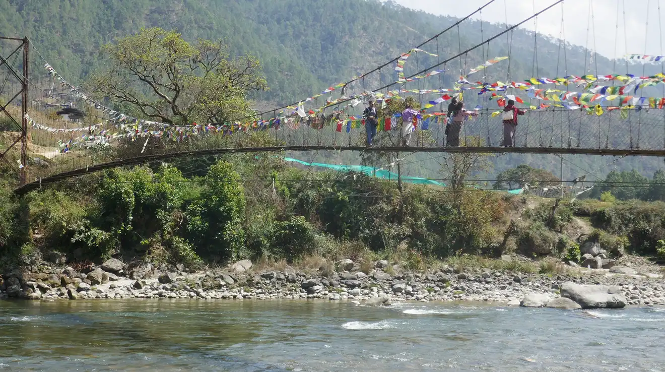 Travelers walk across a long suspension bridge adorned with colorful prayer flags over a flowing river during a scenic Western Bhutan Exploration.
