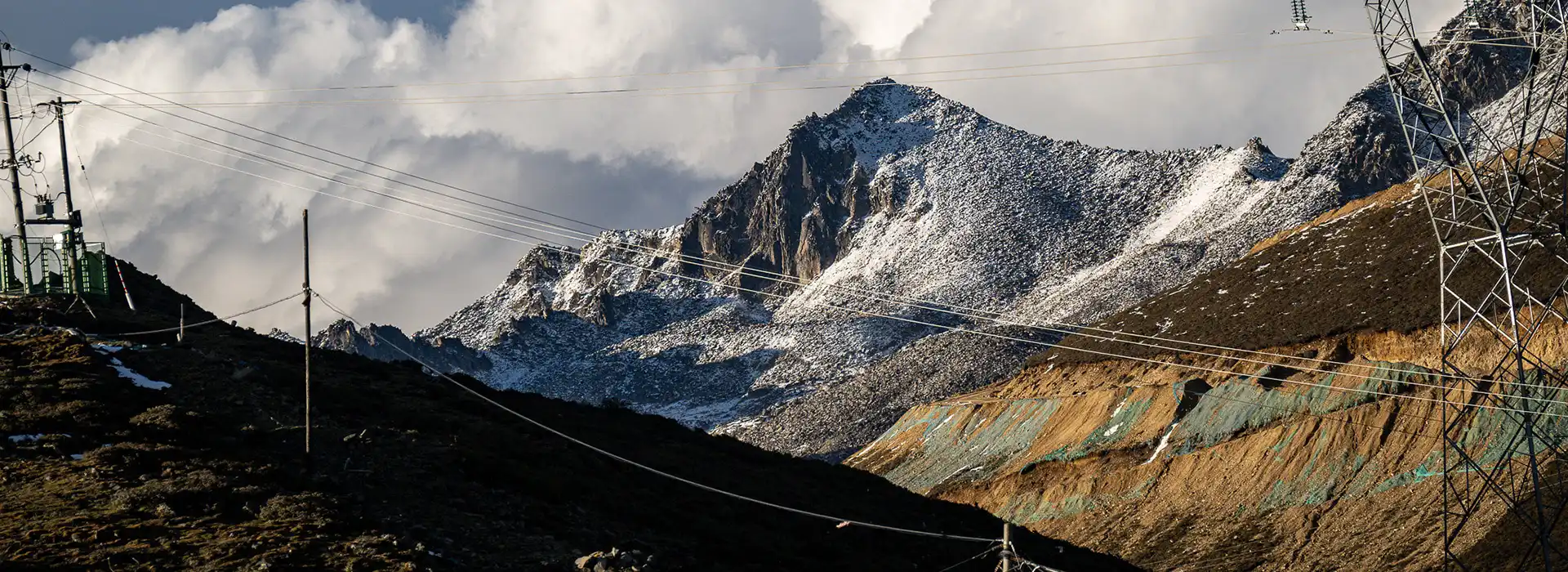 Power lines stretch across a rugged, snow-dusted mountain pass under a dramatic sky. Many travelers crossing these high-altitude border regions book a Tibet tour from India.