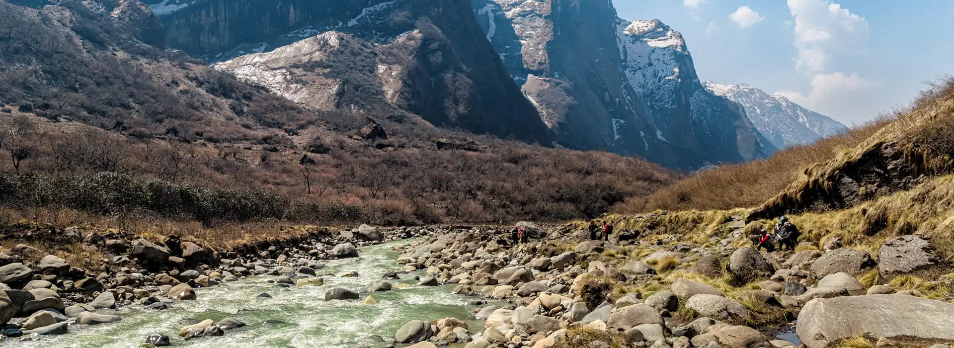 A wide view of a turquoise glacial river flowing through a rocky valley beneath massive, snow-dusted cliffs in the best nature destinations in Nepal.