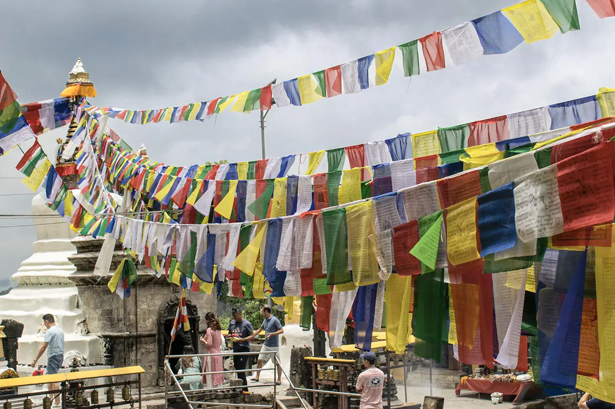 Vibrant Buddhist prayer flags stretch across a courtyard near a white stupa where visitors gather. This spiritual site is a top recommendation in any Kathmandu Travel Guide.