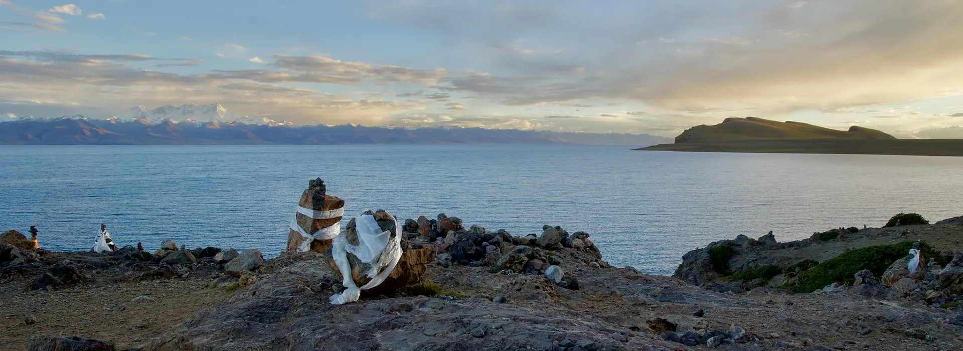 The calm, blue waters of Lake Manasarovar reflecting snow-capped peaks at sunset, with traditional Tibetan cairns on the foreground bank during a Kailash Tour from Lhasa.