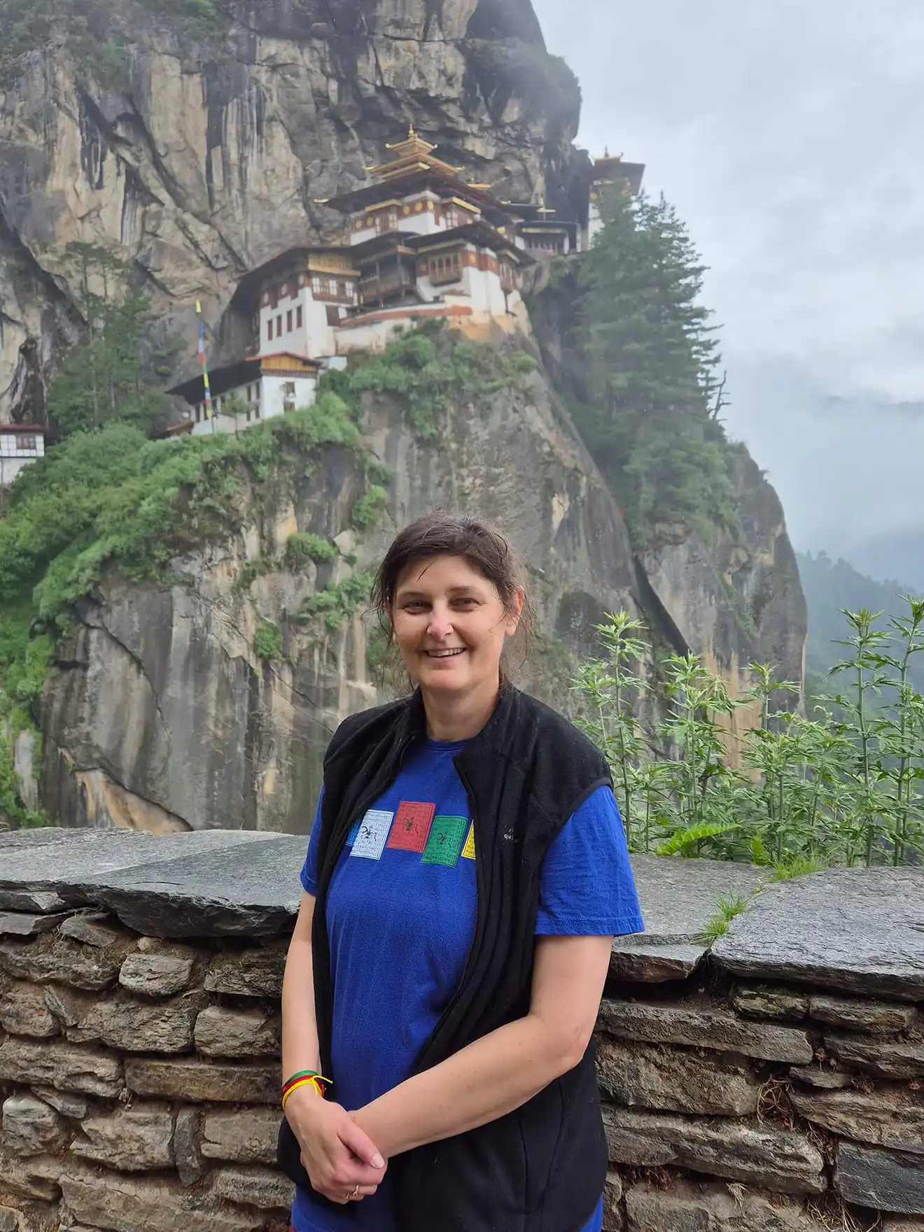 A smiling woman stands before the Tiger’s Nest Monastery, perched on a cliffside. This iconic destination is a major highlight during our Essential Bhutan Travel Tour.