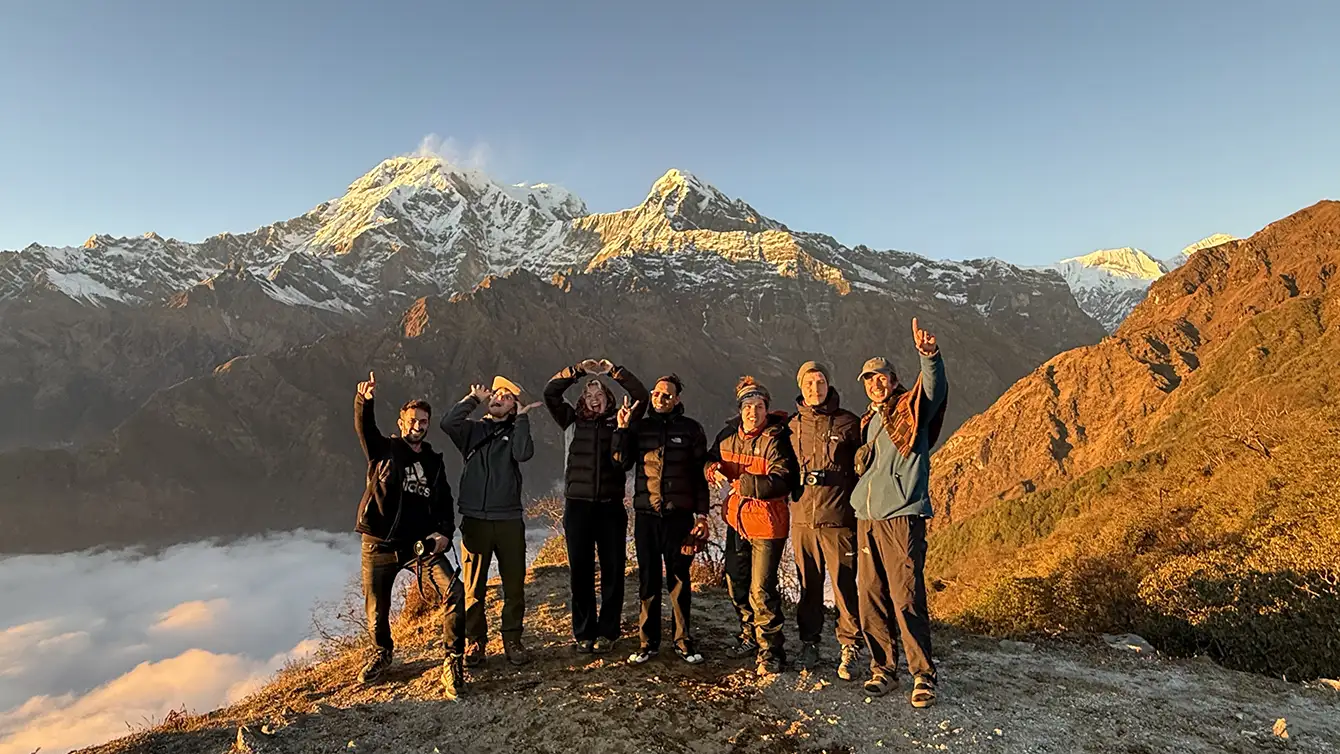 Seven diverse trekkers in winter gear celebrate at a Himalayan viewpoint with massive snow-capped mountains and a sea of clouds, illustrating the destination of a Nepal Hiking Permits Guide trip.